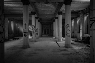 A black and white photo of a dark room with large columns forming a hallway down the center.