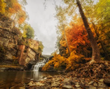 A soft lit image of a waterfall surrounded by vibrant trees with orange and yellow autumn foliage. Nestled into the cliffside to the left of the waterfall is an old brick abandoned hydro-electric station.